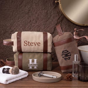 Op de afbeelding: Drie rechthoekige toilettassen met bruine kunstlederen accenten en handvatten. Eén tas is crème met de naam Steve, één is olijfgroen met de naam Hudson en een grote H, en één is bruin met een zwart geborduurd monogram.