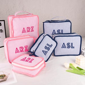 May include: Set of six striped toiletry bags in pink and blue with white stripes. The bags are personalized with the letters "ARZ" and "ASL" in different colors.