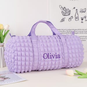 Op de afbeelding: Een lavendelkleurige gewatteerde reistas met een paarse band en de naam "Olivia" geborduurd op de voorkant.