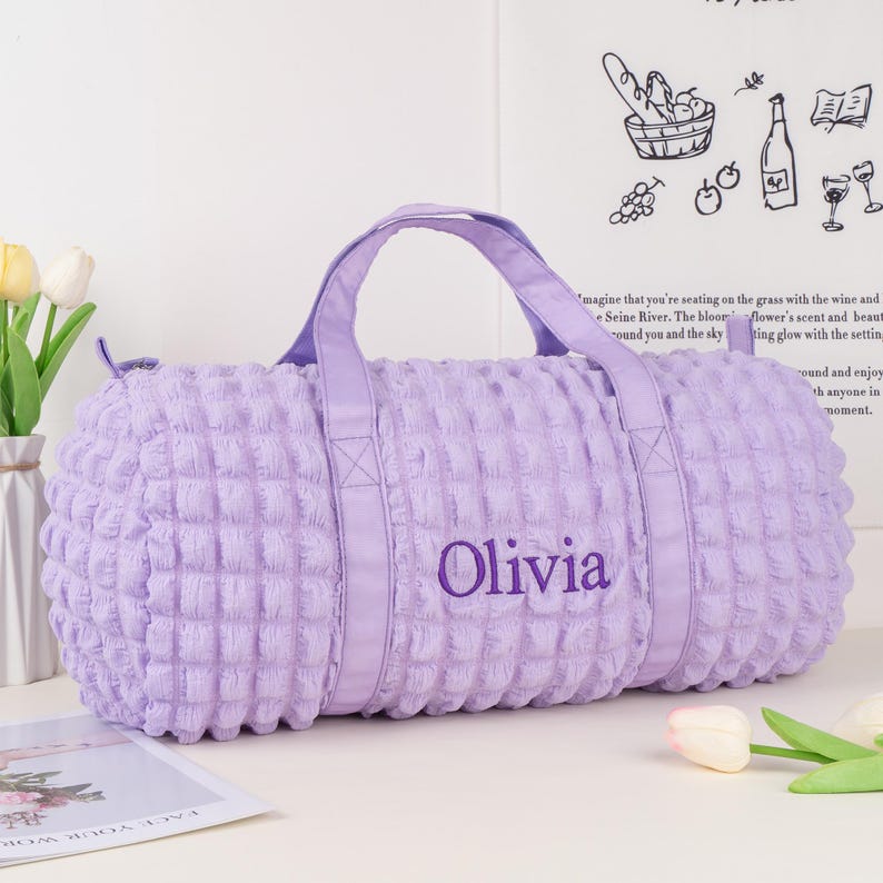 Op de afbeelding: Een lavendelkleurige gewatteerde reistas met een paarse band en de naam "Olivia" geborduurd op de voorkant.