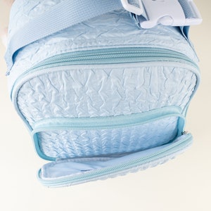 Pu&ograve; includere: Una borsa per pannolini trapuntata di colore azzurro chiaro con una parte superiore con cerniera e una tasca frontale. La borsa ha una tracolla con fibbia in plastica bianca.