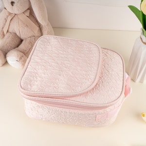 Pu&ograve; includere: Una pochette rosa trapuntata con cerniera, patta superiore e manico. La pochette &egrave; realizzata in un tessuto morbido e testurizzato.