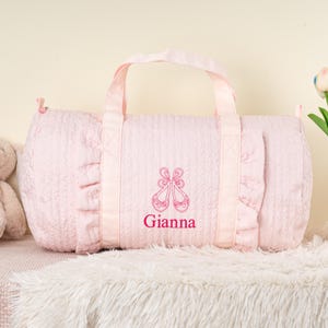 May include: Pink quilted duffel bag with a ruffled trim and a pink embroidered design of ballet shoes and the name "Gianna".