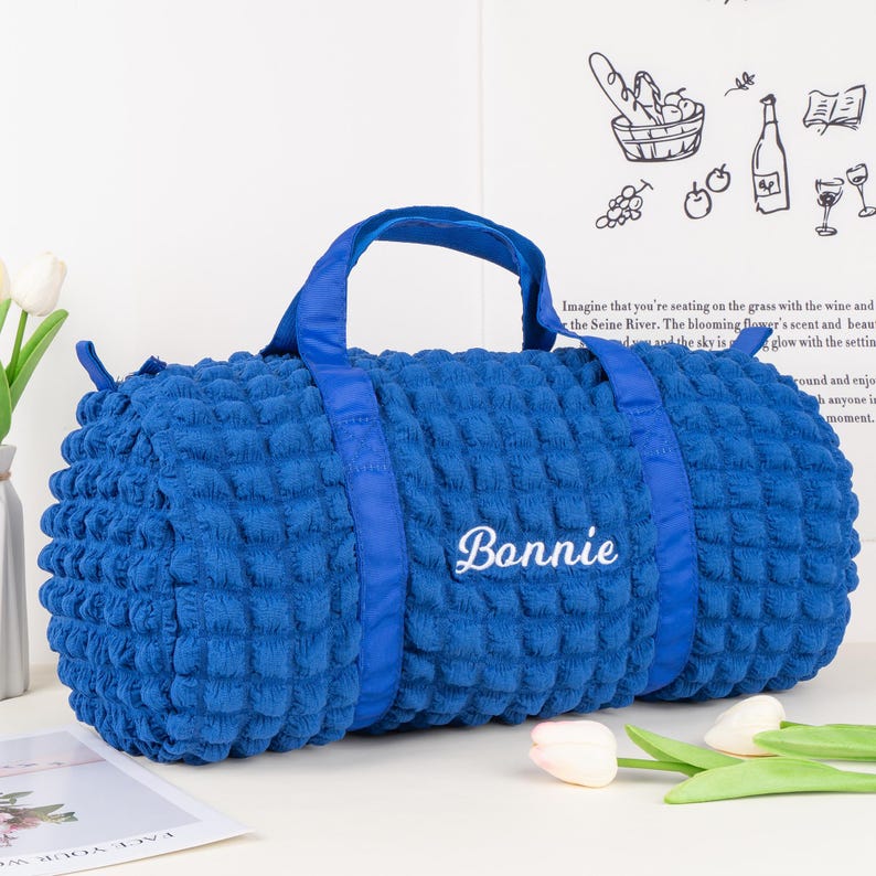 Op de afbeelding: Een blauwe gewatteerde reistas met een blauwe band en de naam "Bonnie" in wit geborduurd op de voorkant.