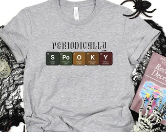 May include: Gray t-shirt with the text "Periodically Spooky" and a periodic table graphic with the elements S, Po, O, K, and Y highlighted. The shirt is on a white background with a skull, spider, and bones.