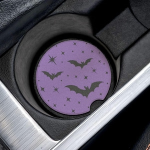 May include: A purple car coaster with a Halloween theme featuring black bats and stars. The coaster is round and fits inside a car's cup holder. The car's interior is black with silver and orange accents.