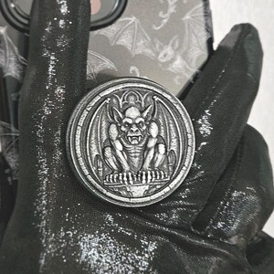 May include: A black smartphone case featuring a repeating white bat pattern and a circular silver-toned phone grip attachment. The grip displays a detailed, embossed gargoyle figure centered within an arched architectural frame design.