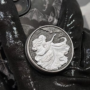 May include: A silver phone grip featuring a detailed relief of a woman in a flowing gown. The grip is attached to a black and gray phone case with a bat design. The phone is held by a black gloved hand.