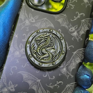 May include: Black phone case with a dragon and bat design. The case has a circular dragon emblem and a pattern of bats. The phone case is held by a person wearing a blue glove.