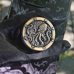 May include: A gold and bronze phone grip featuring an octopus design. The phone case is black with a bat pattern. The grip is attached to a black phone case. The phone case is held by a black glove.