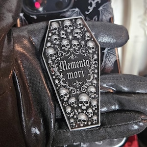 May include: A coffin-shaped pin featuring a black and silver design with numerous small skulls and the text Memento mori in a decorative font. The pin has a silver border and is displayed against a black phone case with a white bat pattern.