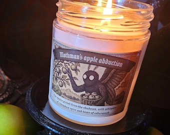 May include: A lit candle in a glass jar with a label that reads "Mothman's apple abduction." The label features a black creature with wings picking apples. Two green apples are in the foreground.
