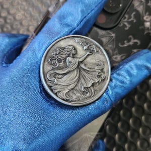May include: A silver-colored round coin featuring a woman with flowing hair reaching for bats. The coin has a detailed relief design and is held against a blue background. The coin is likely a decorative item or collectible.