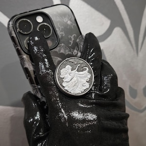 May include: A black and silver phone case with a bat design is shown with a matching phone grip. The phone grip is round with a raised design of a woman in a flowing dress. The phone case and grip are held by a black glove.