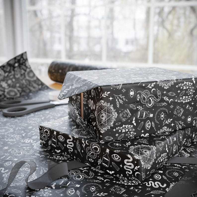 May include: Two gift boxes wrapped in black paper with a white occult-themed pattern. The pattern includes eyes, snakes, potions, and other mystical symbols. A pair of scissors and wrapping paper are visible.