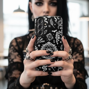 Op de afbeelding: Zwarte telefoonhoes met een wit occulte ontwerp. De hoes heeft een pentagram, kristallen, een slang en andere mystieke symbolen. De hoes wordt voor een persoon gehouden.