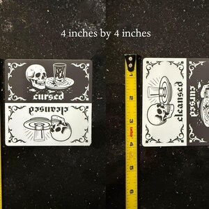 4 by 4 inch magnet. A black and white slim dishwasher magnet with a gothic theme. The top half features a skull, a glass, and the word 'cursed'. The bottom half shows a skull, a glass, and the word 'cleansed'. Ornate flourishes frame the design.
