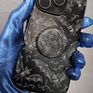 May include: Black phone case with a bat and moon design. The case features a detailed illustration of bats and a textured moon pop socket. The case is held by a blue hand.