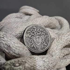 May include: A silver-colored coin featuring a detailed relief of Medusa with snakes for hair. The coin is set against a textured, light gray sculpture of intertwined snakes. The coin's design is intricate and the sculpture has a rough, stone-like appearance.