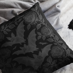 Puede incluir: Cojín cuadrado negro con un patrón repetido de murciélagos y rosas. Los murciélagos se representan con las alas extendidas y las rosas están en plena floración. El diseño tiene una estética monocromática de inspiración gótica.