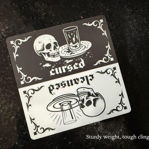Sturdy magnet weight and tough dishwasher magnet cling! A black and white slim dishwasher magnet with a gothic theme. The top half features a skull, a glass, and the word 'cursed'. The bottom half shows a skull, a glass, and the word 'cleansed'. Ornate flourishes frame the design.