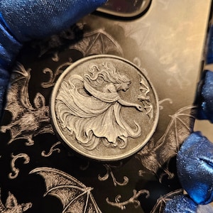 May include: A silver-colored round phone grip featuring a detailed relief of a woman with flowing hair and a flowing gown. The phone case has a black background with a bat pattern.
