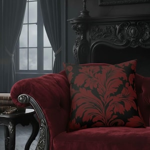 May include: A cushion with a black and red floral design sits on a red velvet sofa. The cushion's pattern is a large, stylised botanical print. The sofa is in a gothic-style room with dark walls and a fireplace.