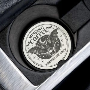 May include: A round, off-white car coaster with a black bat and coffee cup design. The text reads "MISSING COFFEE" and "SUMMON THE BREW" in a circular layout. The coaster sits in a black car cup holder, surrounded by silver and black interior.