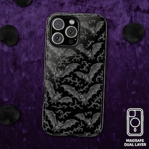 May include: Black phone case with a repeating pattern of bats and swirling vines. The case has a glossy finish and a MagSafe dual-layer design. The bats are detailed with gray shading, creating a gothic aesthetic.