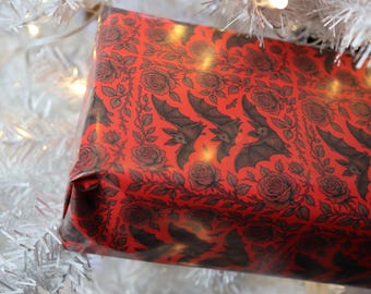 May include: A rectangular gift box wrapped in vibrant red paper featuring a repeating black ink illustration pattern of flying bats and blooming roses. The wrapping paper has a smooth, slightly glossy finish and covers the entire visible surface.