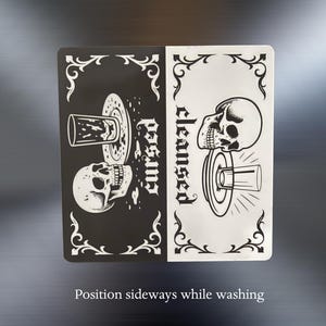 Position sideways while washing. A black and white slim dishwasher magnet with a gothic theme. The top half features a skull, a glass, and the word 'cursed'. The bottom half shows a skull, a glass, and the word 'cleansed'. Ornate flourishes frame the design.