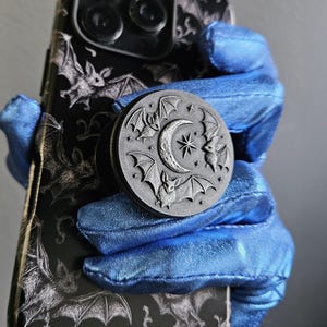 May include: A black phone case with a bat pattern and a round, gray phone grip. The phone grip features a crescent moon, star, and bat design. The phone case is held by blue fabric.