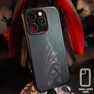 May include: A black dual-layer phone case featuring a detailed, engraved bat design. The case has a matte finish and a cutout for the phone's camera. The image also includes a graphic of the case and the words "DUAL LAYER CASE".