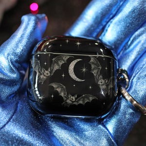 May include: Black AirPod case with a bat and moon design. The case has a glossy finish and a silver keyring attachment. The design includes bats, a crescent moon, and stars.
