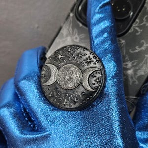May include: A black and silver phone grip featuring a triple moon design with stars. The round grip is attached to a phone case with a dark pattern. The background is a shiny blue fabric.
