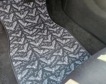 May include: Black car floor mat with a repeating pattern of white bats and decorative swirls. The mat is shaped to fit the car's floor and is placed in front of the pedals. The overall theme is gothic or Halloween.