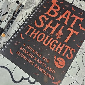 May include: A black spiral-bound journal with the words "BAT SHIT THOUGHTS" in orange lettering. The journal also features the text "A JOURNAL FOR WINGED RANTS AND MIDNIGHT RAMBLINGS". Three Sharpie pens are visible in the image.