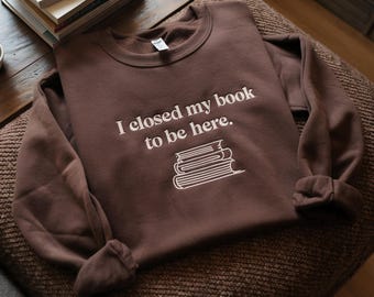 May include: A brown sweatshirt with the phrase "I closed my book to be here" embroidered in white. Below the text is a white outline of a stack of books. The sweatshirt is displayed on a brown surface.