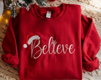 May include: A red sweatshirt with the word "Believe" embroidered in white script, accompanied by a Santa hat and snowflake. The long-sleeved top is displayed on a patterned surface, suggesting a festive theme.
