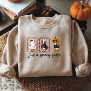 May include: A beige crewneck sweatshirt featuring a graphic of three geese in rectangular frames. The geese are dressed as a ghost, a vampire, and a pumpkin-headed figure. Below the graphic, the text reads, "Just a spooky goose" in black.