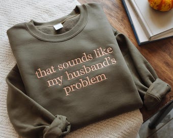 Może przedstawiać: Oliwkowo-zielona bluza z haftowanym napisem "that sounds like my husband's problem" w jasnobrązowym kolorze. Bluza jest złożona na kremowym kocu. Książka i mała dynia są w tle.