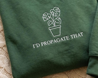 May include: A dark forest green sweatshirt featuring a white line art graphic of a potted monstera plant. Below the plant, the text "I'D PROPAGATE THAT" is embroidered in a clean, white, sans-serif font, centered on the chest area of the garment.