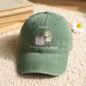 May include: A sage green baseball cap featuring an embroidered graphic of a frog wearing an apron and stirring a silver pot. The text above the frog reads "That's it." and the text below the frog reads "You're going in the SOUP."