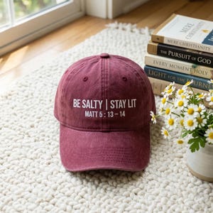 Puede incluir: Gorra de béisbol granate con el texto "BE SALTY | STAY LIT" y "MATT 5:13-14" en blanco. La gorra está sobre una alfombra blanca texturizada. Libros y flores en el fondo.
