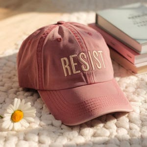 May include: A dusty rose baseball cap with the word "RESIST" embroidered in off-white. The cap has a curved brim and a classic six-panel design. The hat is sitting on a white, textured surface.