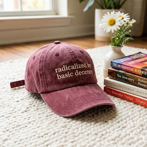 May include: A maroon baseball cap with the phrase "radicalized by basic decency" embroidered in white. The cap is sitting on a white rug, with books and flowers in the background.