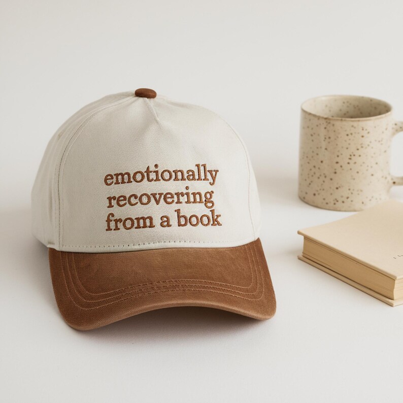 May include: A baseball cap with a white crown and a brown brim. The cap has the embroidered text "emotionally recovering from a book" in brown. A ceramic mug and a closed book are in the background.
