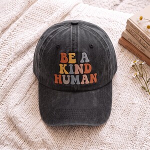 May include: A charcoal grey baseball cap with a distressed, washed finish. The front features the embroidered text BE A KIND HUMAN in a retro, multi-coloured font with shades of orange, yellow, and light blue. The cap has a structured crown.