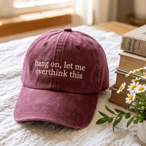 May include: A maroon baseball cap with a structured crown and a curved brim. The front of the cap features the embroidered white text "hang on, let me overthink this" in a simple, lowercase font. The fabric has a slightly washed, textured appearance.