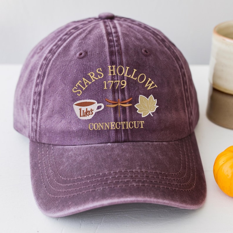 May include: A purple baseball cap with the words "STARS HOLLOW 1779 CONNECTICUT" embroidered in gold. The design includes a coffee cup, dragonfly, and maple leaf. The cap has a curved brim and adjustable strap.